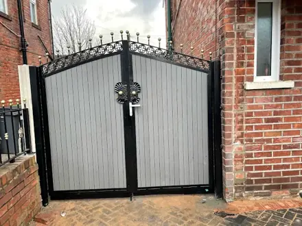 Grey composite double gates in a black frame with arched scrollwork and gold finials, beside a red-brick house
