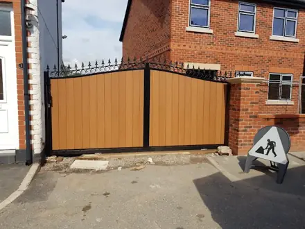Wood-effect double driveway gates in a black frame with scrollwork and spear tops, between a white-painted wall and a brick pillar, with a roadworks sign beside them
